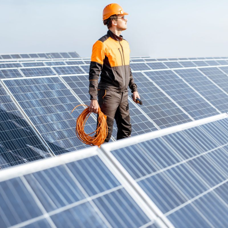 Well-equipped worker in protective orange clothing servicing solar panels on a photovoltaic rooftop plant. Concept of maintenance and installation of solar stations